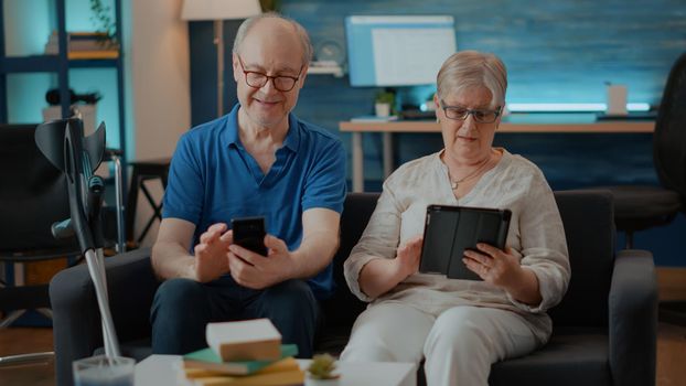 Modern couple using smartphone and digital tablet at home. Old people with physical disability looking at mobile phone and gadget to have fun with technology in living room. Senior family