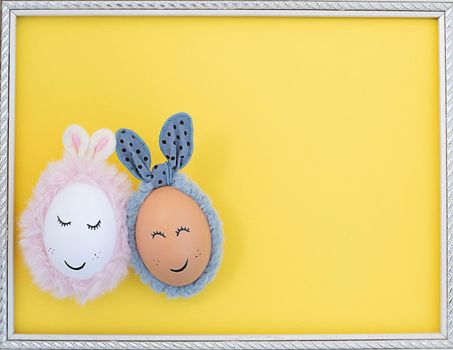 Two Easter eggs with painted eyes and rabbit ears on a yellow background in a white frame. close up