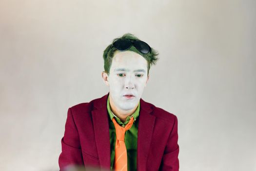Person in bordo suit, shirt with yellow stripes tie looks down. Actor with white makeup on face, color eyes and green hair. Paint of clown or mime. Theater concept. Black glasses on head.