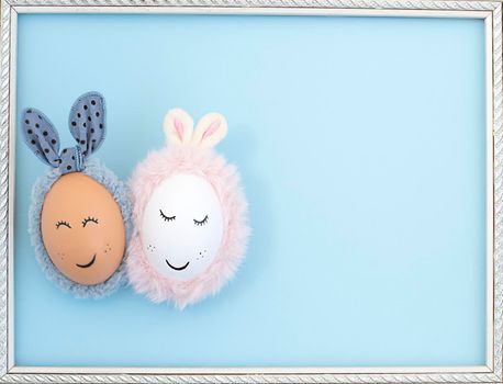Two Easter eggs with painted eyes and rabbit ears on a blue background in a white frame. close up