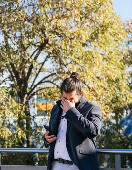 young businessman with long hair and beard, brown, laments because he receives bad news from his smartphone. He is outside near his offices during his work break.