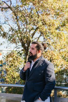 Young businessman with long hair and beard standing minding his own business during his work break, on the street next to the offices. Vertical photo on a sunny and clear day.