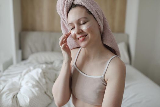 beauty, skin care, spa concept, smiling young woman with a towel on head apply cream on face skin