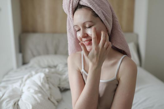 smiling young woman with a towel on head apply cream on face skin