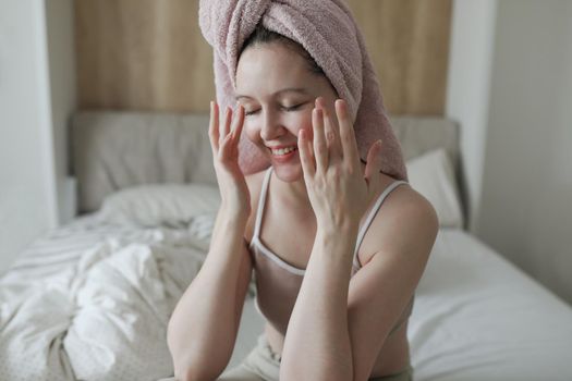 beauty, skin care, spa concept, smiling young woman with a towel on head apply cream on face skin