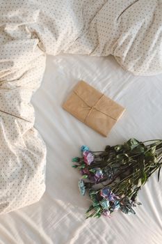 Bouquet of flowers and a gift covered in paper on crumpled white bed sheets.
