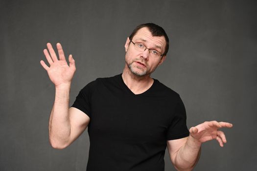 Adult man in a black t-shirt and glasses talking with emotions to the camera on a gray background