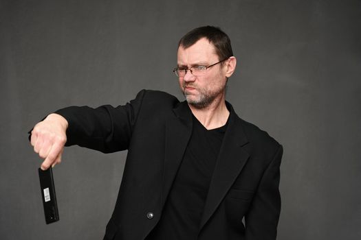 Portrait of an adult man in a jacket with glasses with displeasure looks at the phone in his hands on a gray background