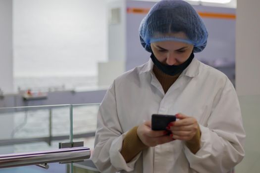 A quality inspector or food specialist is on site. An employee in a white coat and hat at an industrial facility looks into a smartphone. Confectionery factory worker standing in a white coat