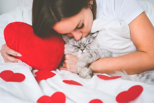Woman lying on the bed with red hearts and kissing her lovely cat. Love for pets. Valentine's day concept. High quality photo
