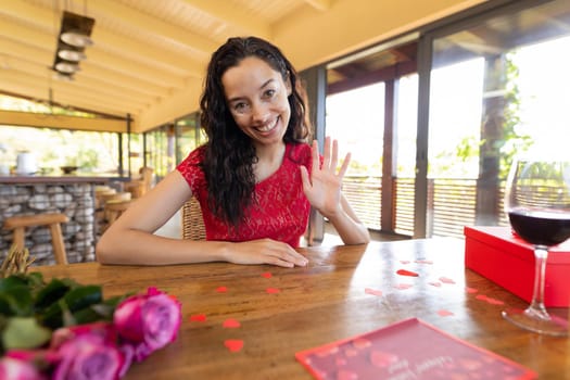 Portrait of smiling biracial young woman with wine and gift waving hand in restaurant. unaltered, online dating, vitiligo and distant valentine day celebration.