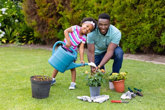 Portrait of smiling african american girl watering plants by father kneeling in garden. family, love and togetherness concept, unaltered.