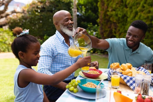 Smiling african american man serving juice for daughter by senior man at dining table in backyard. family, love and togetherness concept, unaltered.