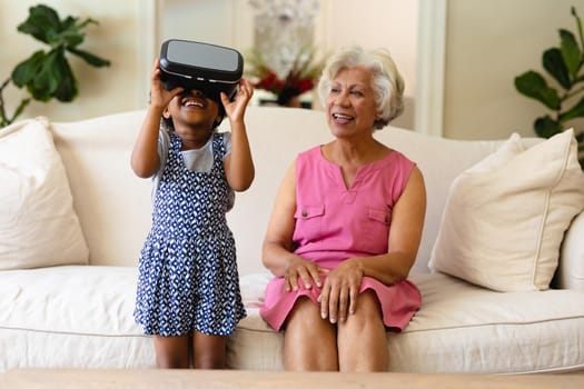 African american girl wearing vr headset and grandmother smiling sitting on the couch. virtual reality and futuristic technology concept, unaltered.