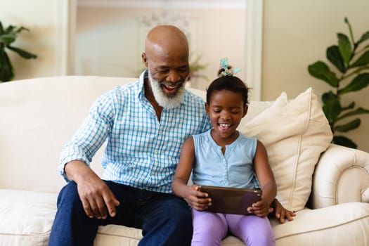 Smiling african american senior man with granddaughter using digital tablet while sitting on sofa. unaltered, wireless technology, family, lifestyle, leisure activity and domestic life concept.