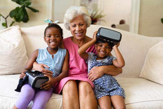 African american grandmother and her two granddaughters with vr headsets sitting on couch at home. virtual reality and futuristic technology concept, unaltered.