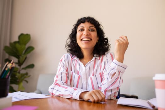 Portrait of smiling young biracial businesswoman sitting at desk in office. happy female professional at workplace, unaltered.