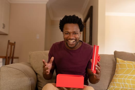 Portrait of cheerful excited african american man opening valentine's present box sitting at home. lifestyle, celebration and surprise, unaltered.