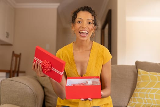 Portrait of cheerful excited biracial woman opening valentine's present box sitting on sofa at home. lifestyle, celebration and surprise, unaltered.