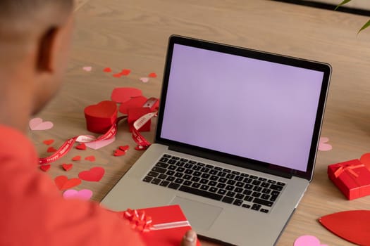 African american man with valentine's gift on laptop video call with blank screen showing copy space. technology, bonding and surprise, unaltered.