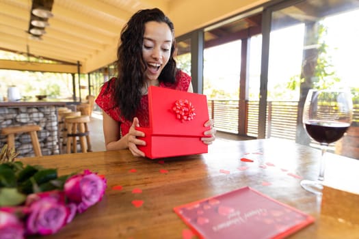 Surprised biracial young woman opening gift box at table in restaurant. unaltered, online dating, vitiligo and distant valentine day celebration.