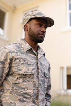 African american army man wearing camouflage uniform and cap looking away against house. identity and patriotism, unaltered.