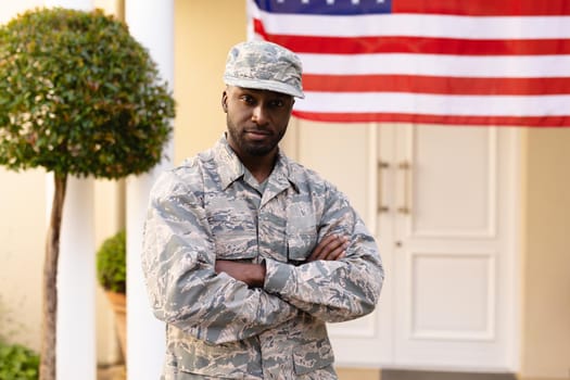 Portrait of confident african american army man in uniform with arms crossed against flag on house. identity and patriotism, unaltered.