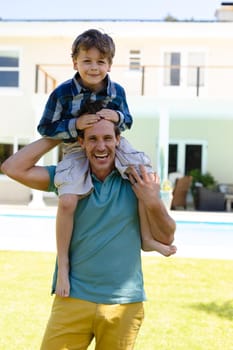 Portrait of caucasian father carrying his son on his shoulders while standing in the garden. family, love and togetherness concept, unaltered.