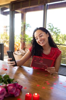 Smiling biracial young woman with smart phone reading greeting card in restaurant. unaltered, online dating, video call, vitiligo and distant valentine day celebration.