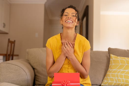 Portrait of happy young biracial woman sitting with valentine's present box on sofa at home. lifestyle and surprise, unaltered.