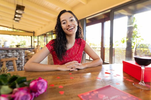 Portrait of smiling biracial young woman with wine and gift at table in restaurant. unaltered, online dating, vitiligo and distant valentine day celebration.