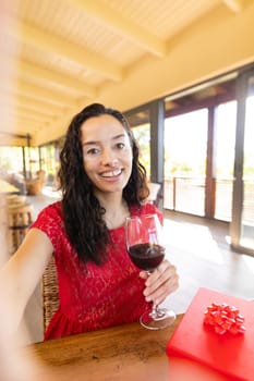 Portrait of happy biracial young woman with red wine video calling in restaurant. unaltered, online dating, vitiligo and distant valentine day celebration.