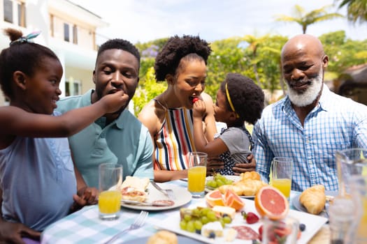 African american girls feeding parents at dining table in backyard during brunch. family, love and togetherness concept, unaltered.