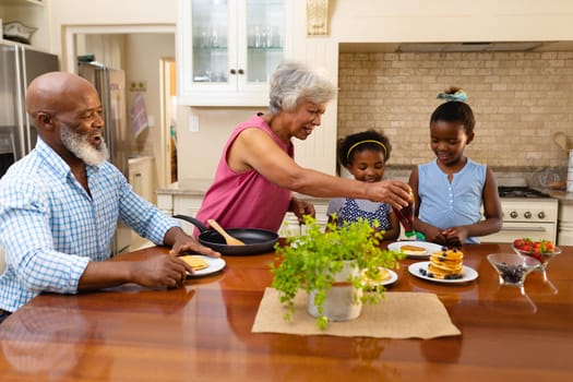 African american grandmother putting maple syrup over the pancake of her granddaughters in kitchen. family, love and togetherness concept, unaltered.