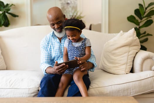 African american grandfather and granddaughter using digital tablet sitting on couch at home. family, love and technology concept, unaltered.