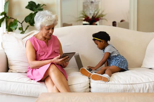 African american grandmother using digital tablet and her granddaughter using laptop on couch. family, love and technology concept, unaltered.