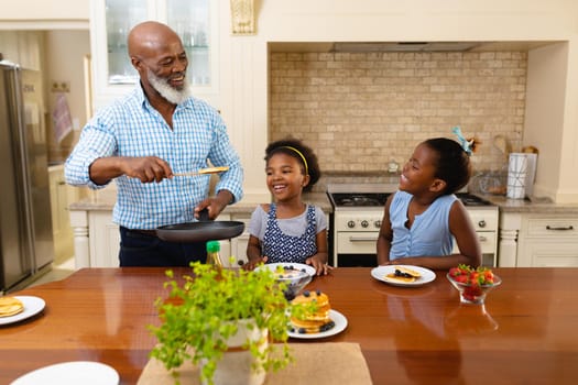 African american grandfather serving breakfast for his two granddaughters in the kitchen at home. family, love and togetherness concept, unaltered.