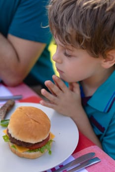 Caucasian boy with hands clasped and eyes closed praying by burger on table in garden. people, food and spirituality concept, unaltered.
