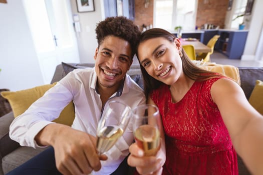 Portrait of smiling young biracial couple with champagne flutes taking selfie at home during party. lifestyle, celebration and memories.