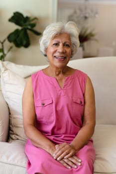 Portrait of smiling biracial mature woman with gray hair sitting on sofa at home. unaltered, lifestyle, leisure activity and domestic life concept.