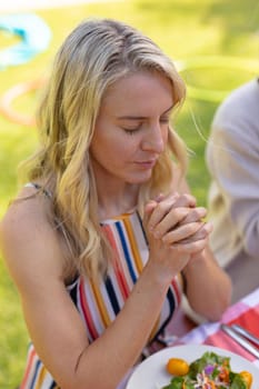 Blond caucasian woman with hands clasped and eyes closed praying at table. people and spirituality concept, unaltered.