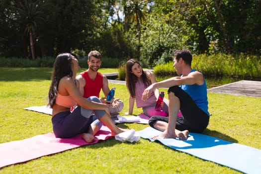 Happy men and women talking while sitting on exercise mats after workout in park. yoga, healthy lifestyle and friendship.