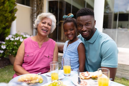Portrait of smiling african american girl with father and grandmother at dining table in backyard. family, love and togetherness concept, unaltered.