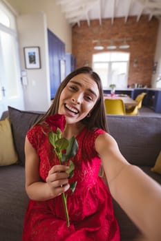 Portrait of happy beautiful biracial woman holding rose bouquet during virtual date at home. lifestyle and virtuality.