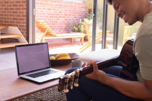 Smiling african american young man playing guitar while using laptop with blank space, copy space. unaltered, lifestyle, leisure activity, music, food, wireless technology and domestic life concept.