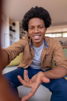 Portrait of smiling african american young man gesturing during video call while sitting at home. unaltered, video call, lifestyle, leisure activity and domestic life concept.