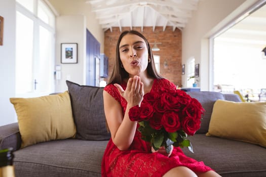 Portrait of biracial woman holding rose bouquet blowing air kiss during virtual date at home. lifestyle and virtuality.