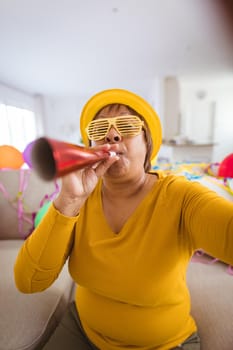 Carefree senior woman in shutter glasses blowing party horn during video call at home. lifestyle, celebration, young at heart and communication.