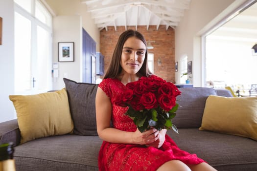 Portrait of smiling attractive biracial woman holding rose bouquet during virtual date at home. lifestyle and virtuality.