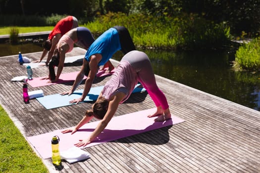 Full length of men and women practicing yoga on floorboard by pond in public park. healthy lifestyle and fitness.